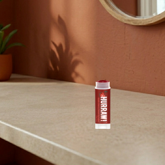 Hurraw! black cherry tinted lip balm on a beige surface with a brown wall and plant in the background