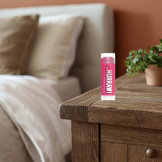 Hurraw watermelon lip balm on a wooden nightstand with a blurred bedroom background