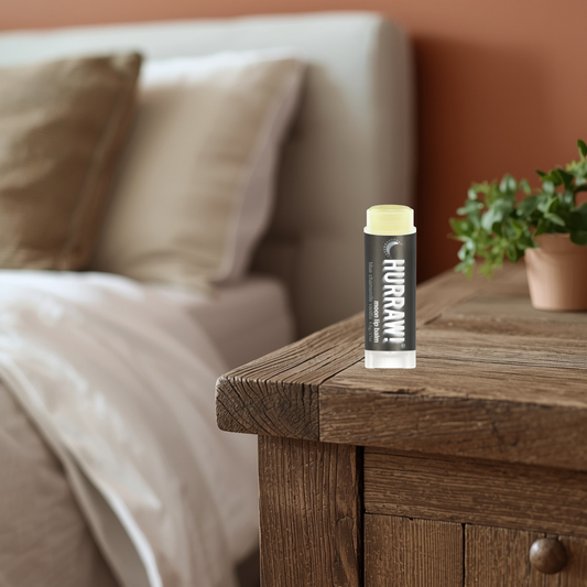 Hurraw moon lip balm on a wooden nightstand with a bed and plant in the background